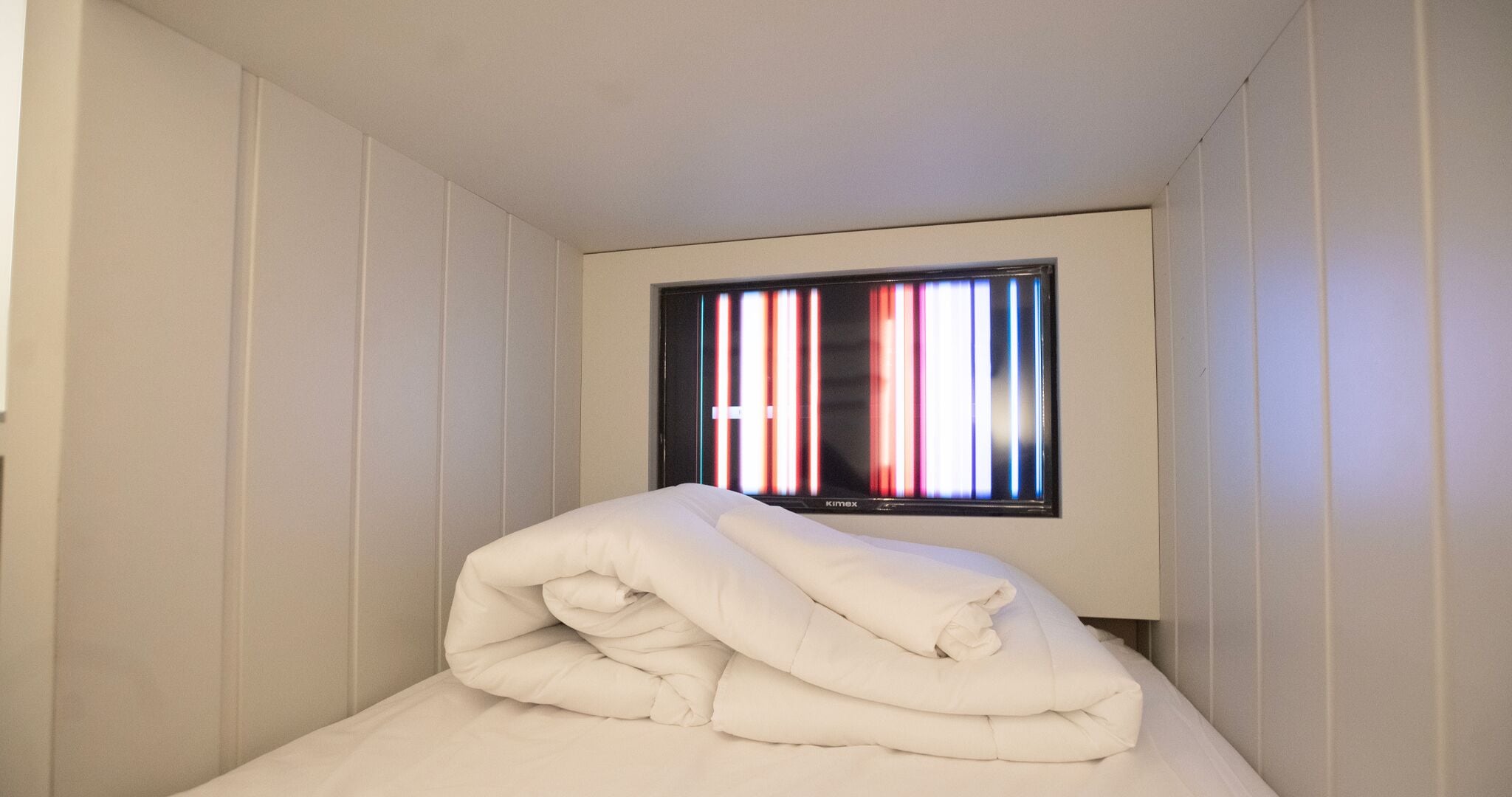 Een kleine, witte kamer met een bed, een raam met verticale zonwering en een aan de muur bevestigde televisie.