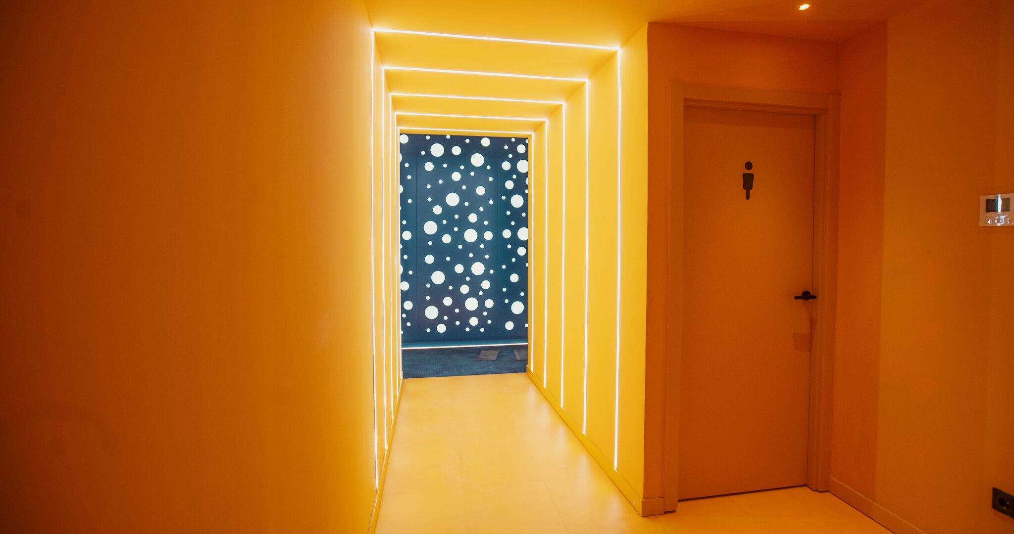 A hallway with a blue wall featuring white polka dots, leading to a door with a light switch and a power outlet.