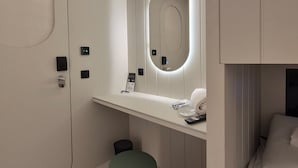 Una camera d'albergo moderna con un comò bianco, uno specchio con luce circolare, un piccolo cestino e un letto con biancheria da letto bianca.