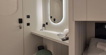 Una habitación de hotel moderna con un tocador blanco, un espejo con luz circular, un cesto de basura pequeño y una cama con ropa blanca.