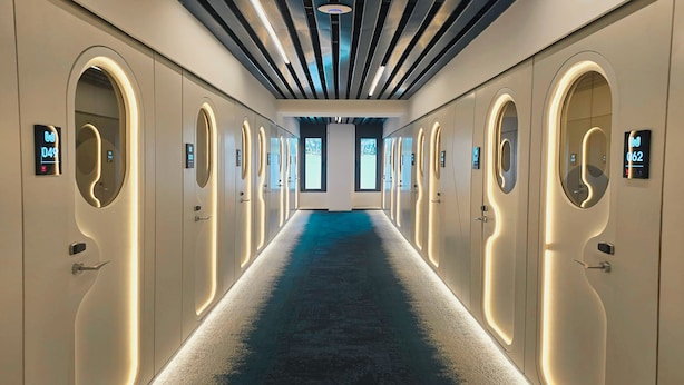 Un corridoio d'albergo con tappeto blu, porte ad arco e numeri illuminati che indicano le camere.