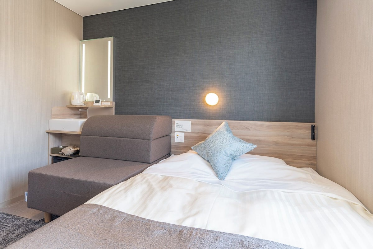 Une chambre d’hôtel moderne équipée d’un lit, d’un petit canapé, d’une table de chevet, d’un miroir et d’une lampe fixée au mur.