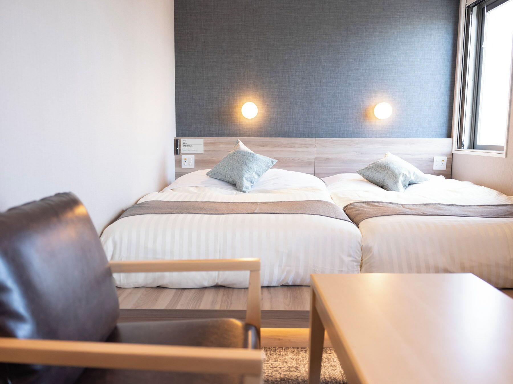 A double bed with two pillows, a bedside table, a chair, and a window with natural light.