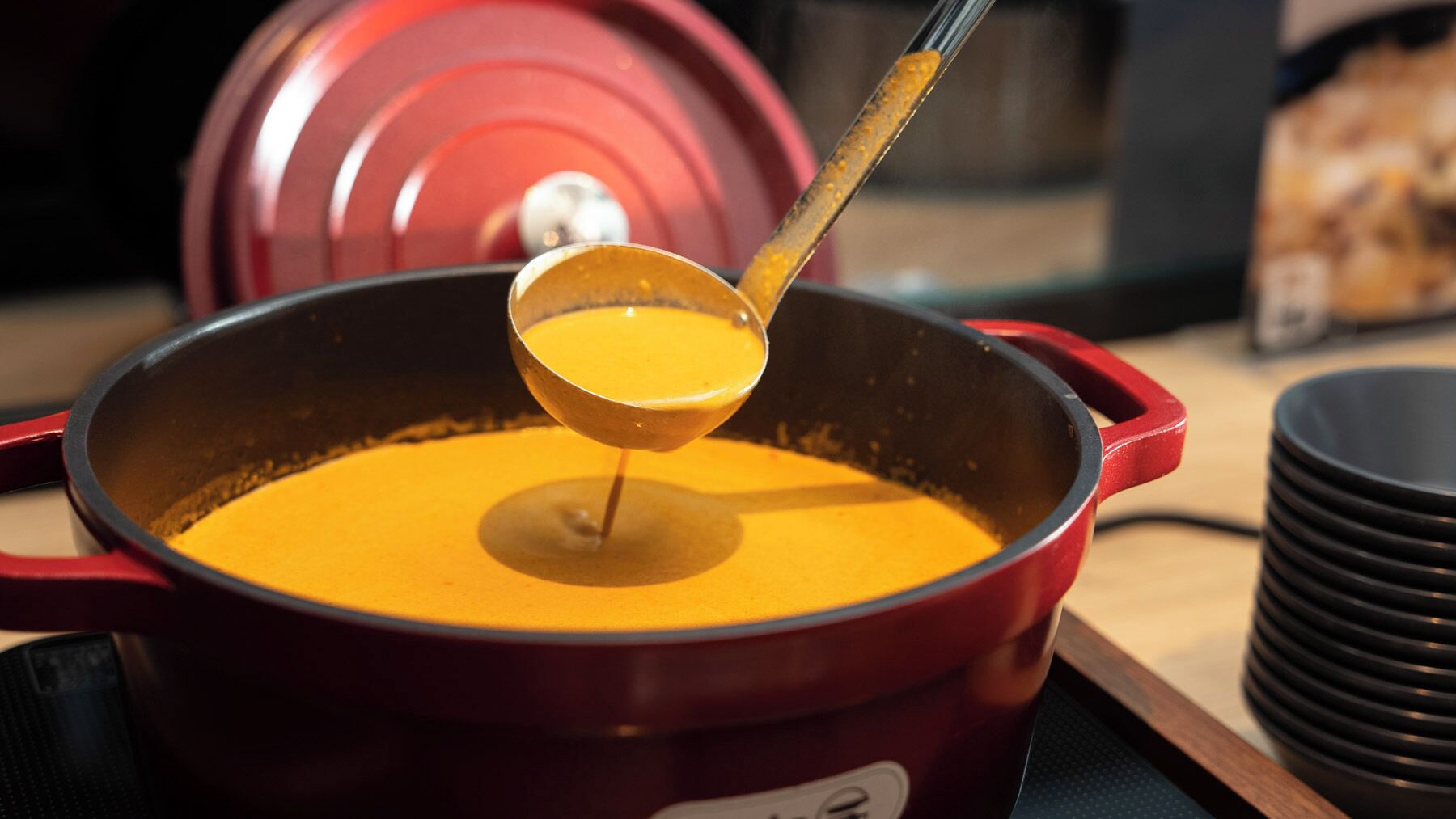 A red pot with a ladle lifting a yellow liquid, likely soup, from a pot.
