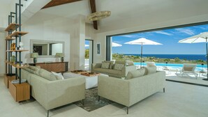 A modern living room with a large sofa, coffee table, and side tables, overlooking a pool and ocean view.