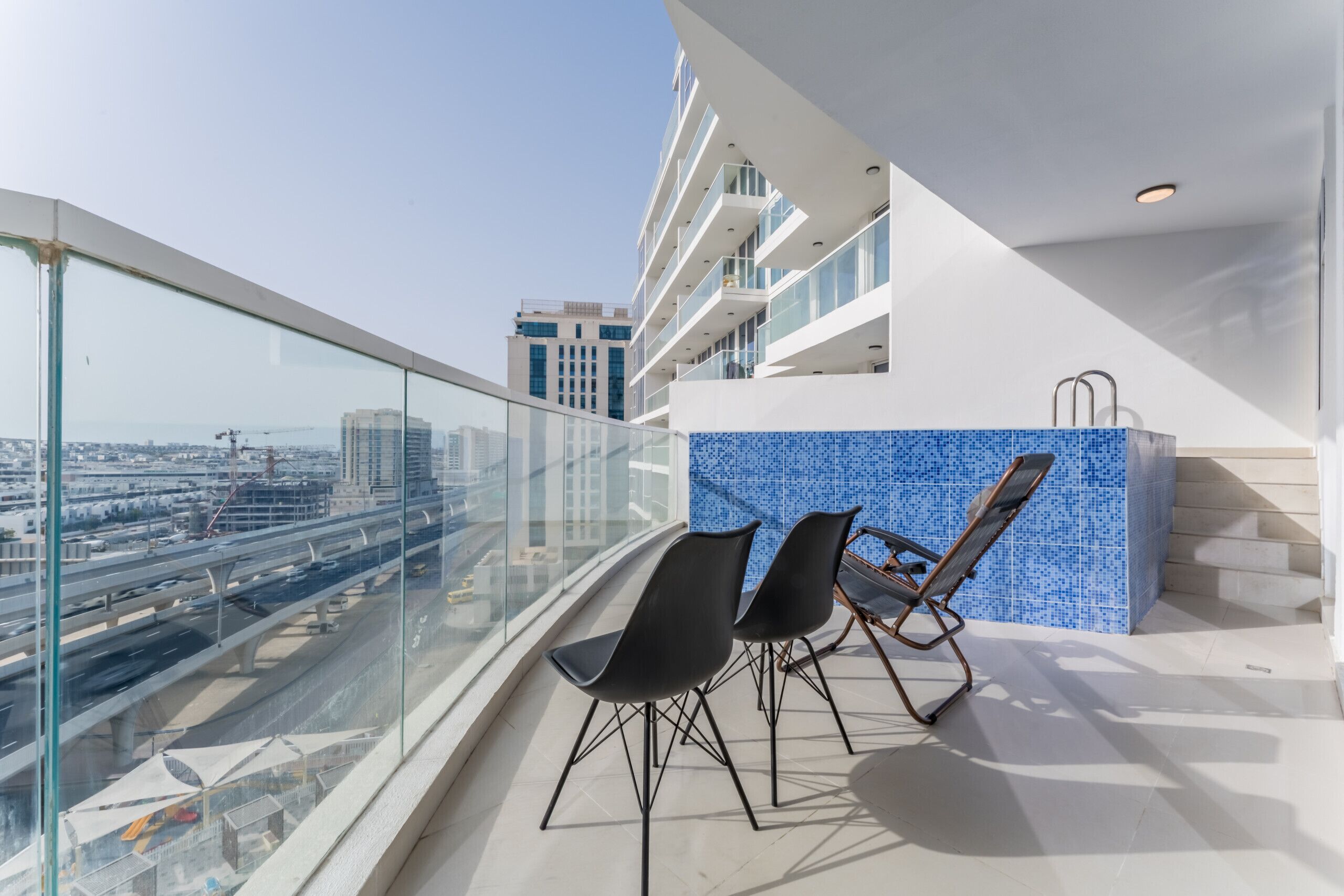 A balcony with a glass railing, two black chairs, and a blue-tiled wall.