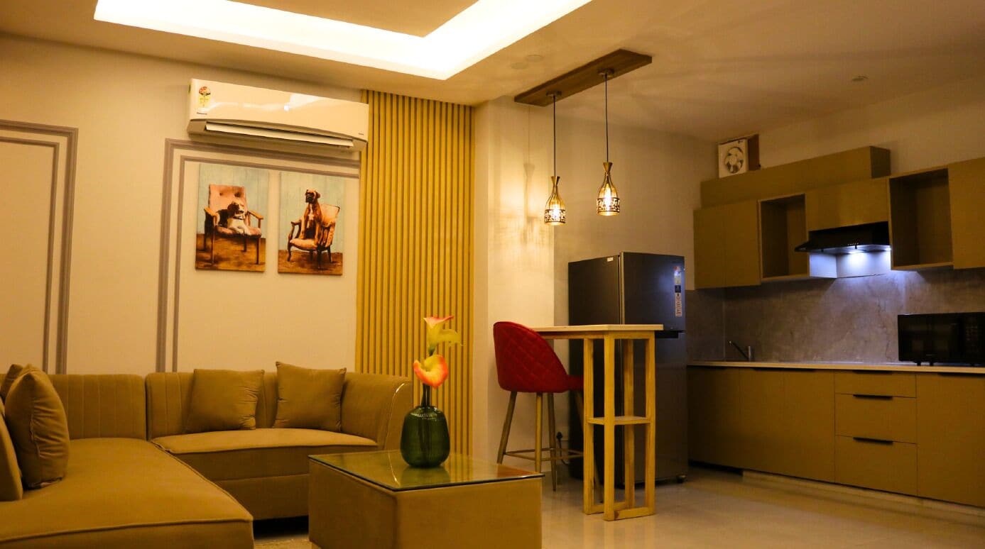 A modern living room with a sofa, a glass coffee table, and a red chair. There is a kitchen area with a refrigerator and a microwave.
