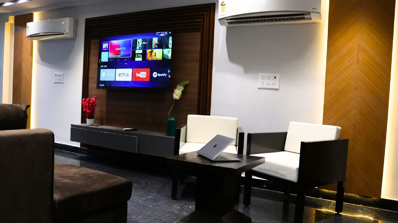 Uma área de espera moderna com televisão de tela plana exibindo aplicativos de streaming, assentos confortáveis, uma mesa de centro com um laptop e um ar-condicionado.