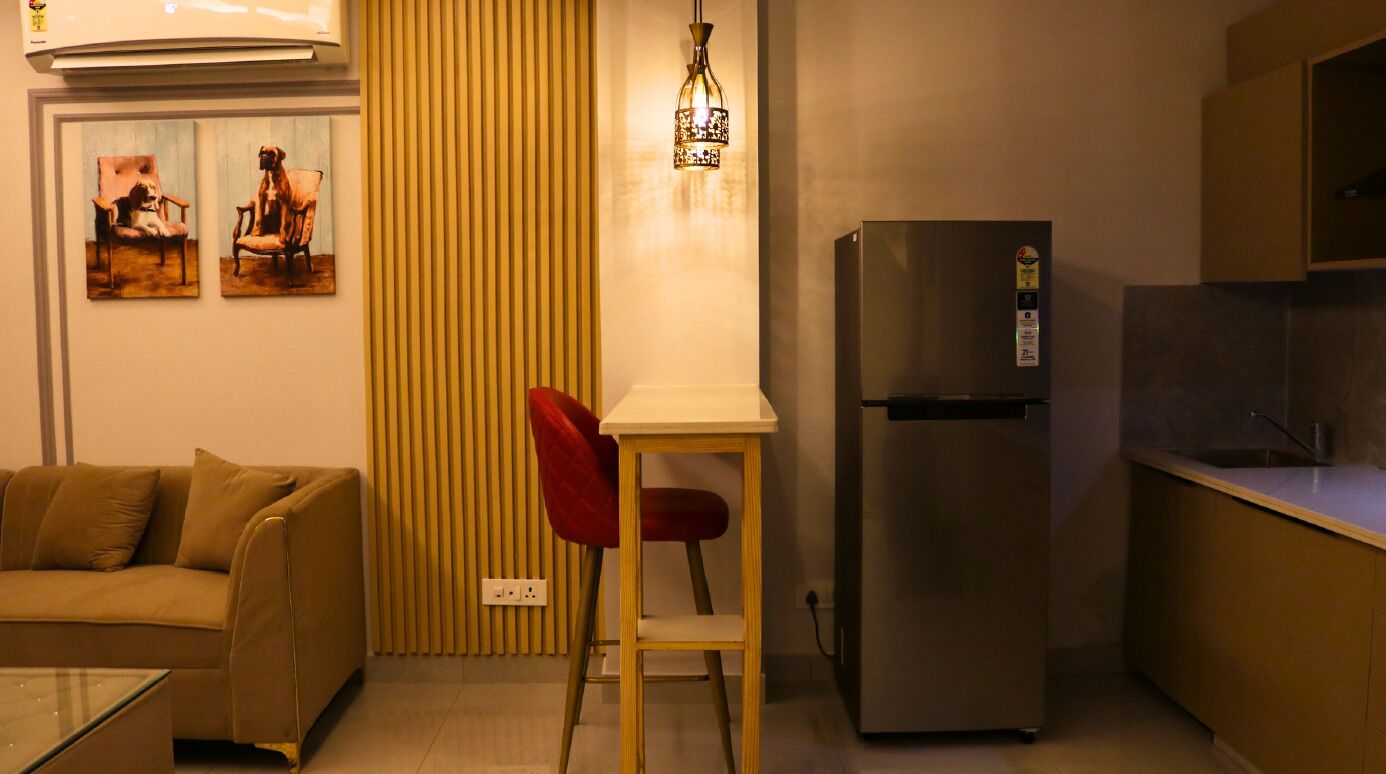 A compact living space with a sofa, a dining table with a red chair, a refrigerator, and a wall-mounted light fixture.