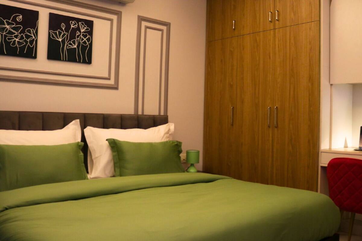 Een slaapkamer met een bed, groene kussens, een bureau met een laptop, een rode stoel en een houten garderobe.