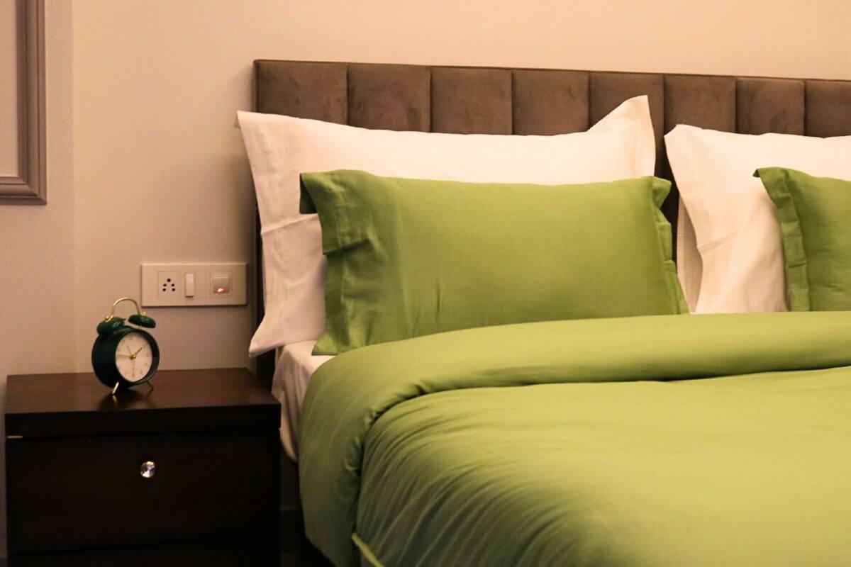 Een net opgemaakt bed met groen-witte beddengoed, een bruin hoofdbord en een klein nachtkastje met een groene wekker.