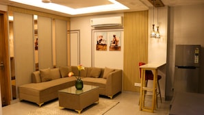 A living room with a sectional sofa, a glass coffee table with a vase, a dining area with a small table and chairs, and a kitchen area with a refrigerator. - Fuji Ryokan Corporate Stays (Gurugram)