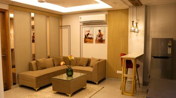 Una sala de estar con un sofá modular, una mesa de centro de vidrio con un florero, un comedor con una pequeña mesa y sillas, y una zona de cocina con un refrigerador.