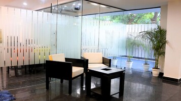 Un área de recepción moderna con divisiones de vidrio, un espacio de descanso con sillas y una mesa, y plantas en macetas.