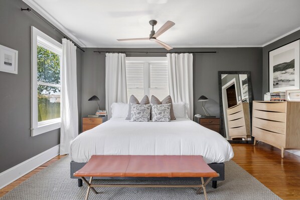 Un dormitorio con cama, una banca, ventilador de techo, cómoda y ventana con cortinas.