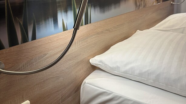 Ein Hotelzimmer mit einem Bett, einem Nachttisch, einer Wandlampe und einem großen Wandbild mit einem See und Bäumen.