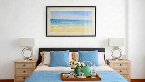 Ein ordentlich eingerichtetes Schlafzimmer mit einem Bett, zwei Nachttischen, einem gerahmten Bild von einem Strand und einer Ablage mit Teeutensilien.