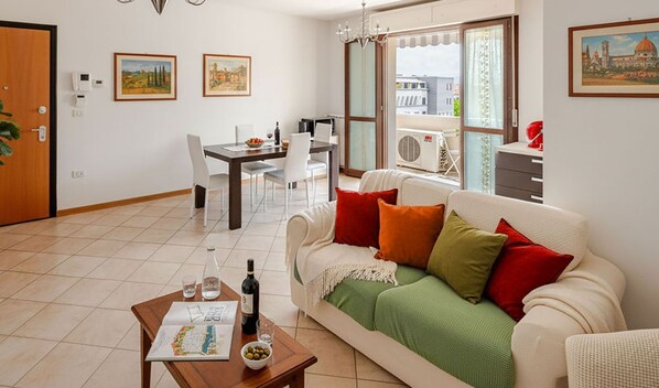 Una sala de estar con comedor, un sofá con cojines, una mesa de centro con una botella y vasos, y un cuadro en la pared.