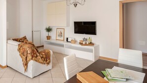 Ein modernes Wohnzimmer mit einem weißen Sofa, einem Esstisch mit Büchern und einem Notizbuch, einem Fernseher an der Wand und einer kleinen Pflanze auf einem Regal.