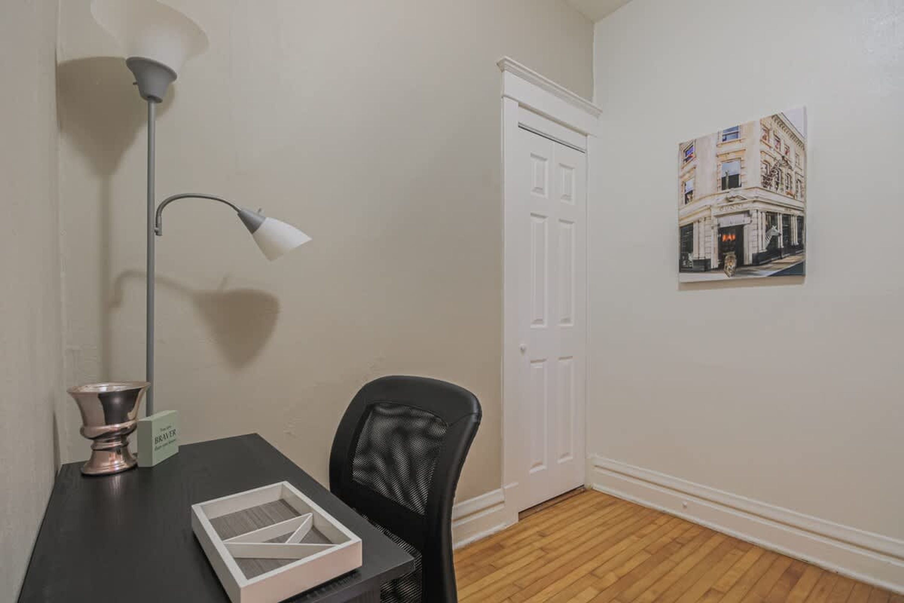 A room with a wooden floor, a black office chair, a desk with a lamp and a decorative bowl, a white door, and a framed picture on the wall.