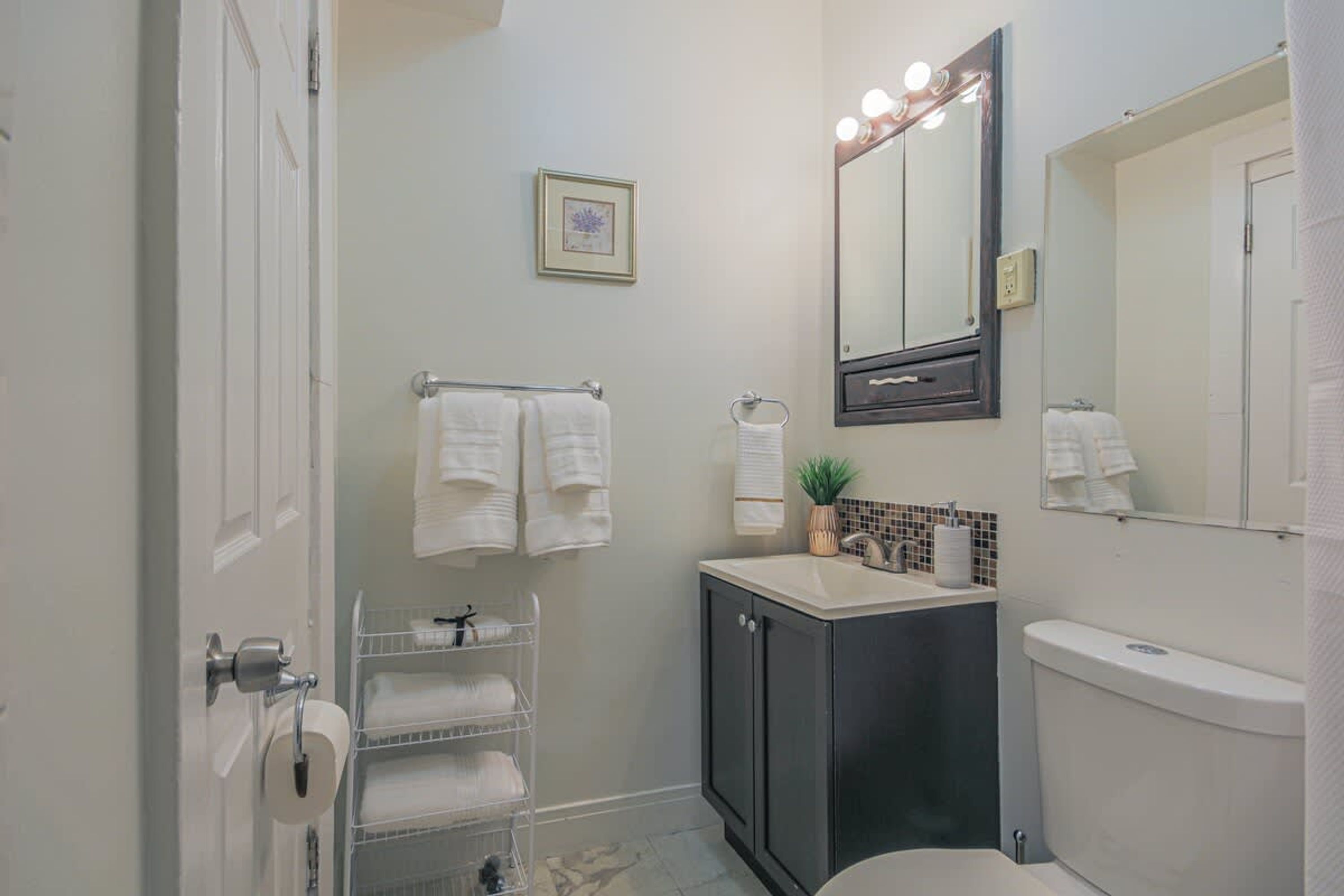 A bathroom with a toilet, sink, and mirror, with towels on a rack and a framed picture on the wall.