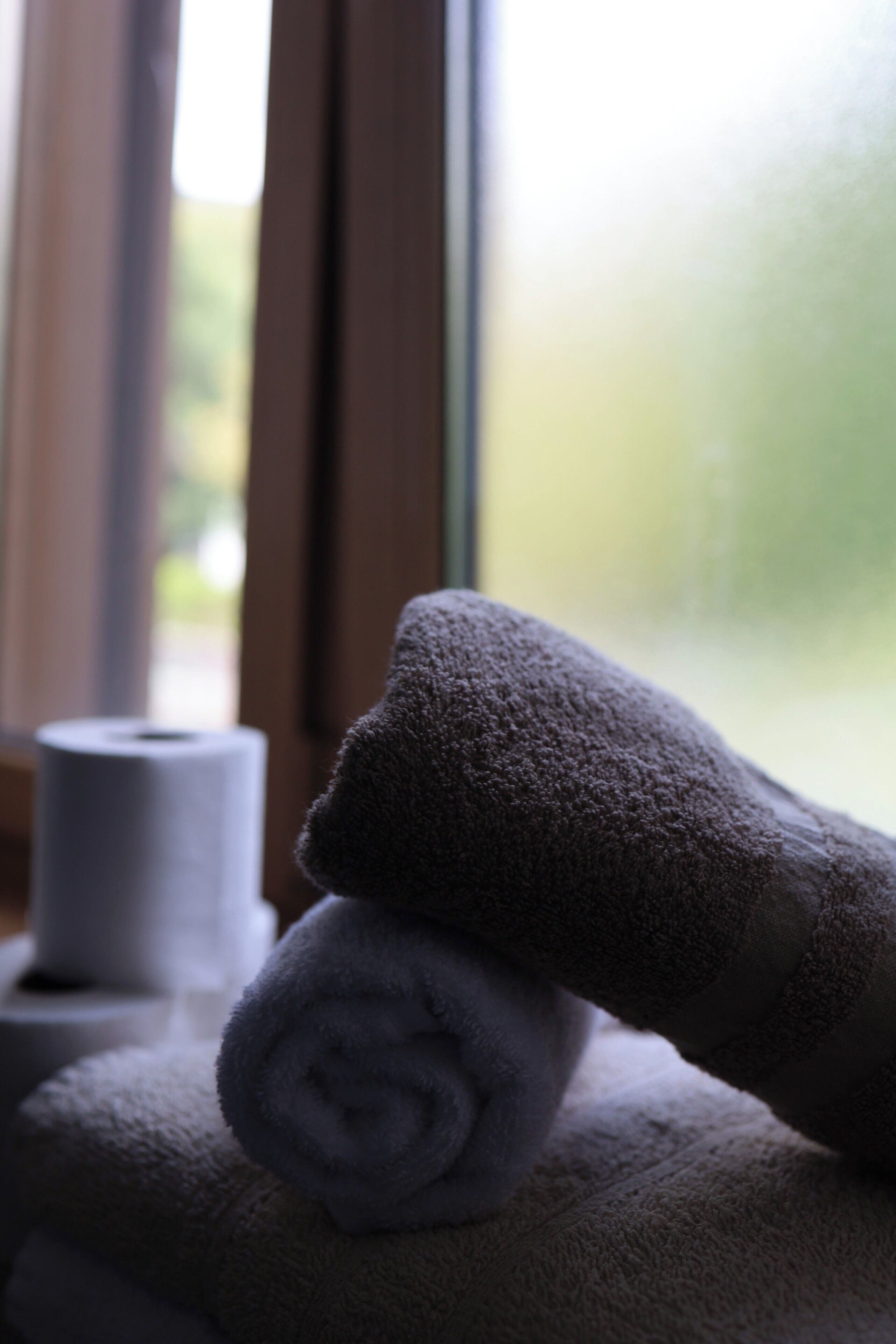 A rolled towel and a roll of toilet paper on a surface near a window.