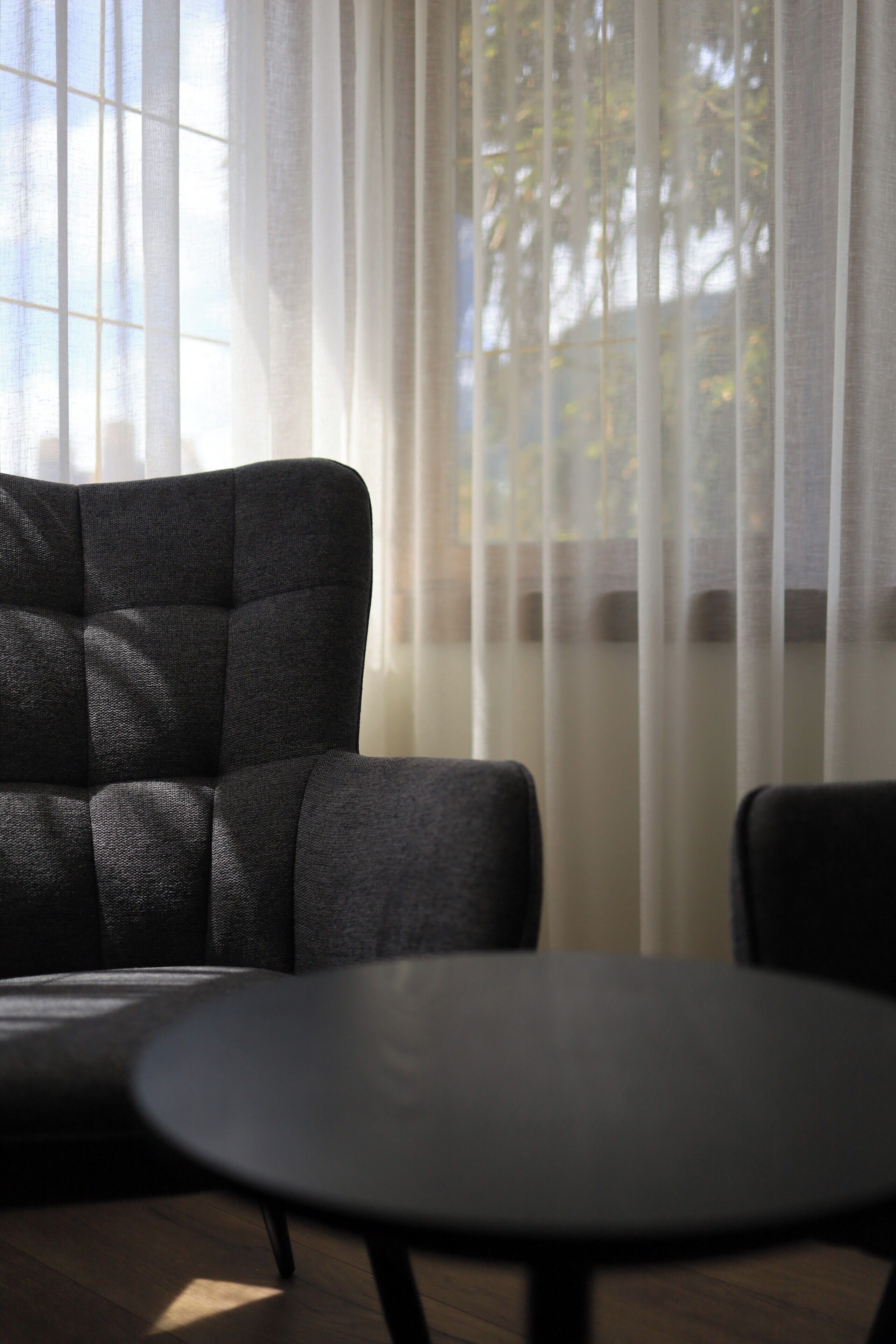 A room with a dark-colored sofa and a round table, with sheer curtains and a window in the background.