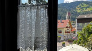 Uma janela com cortinas de renda, vista de uma cidade com edifícios e montanhas, e uma mesa com guarda-sol do lado de fora.