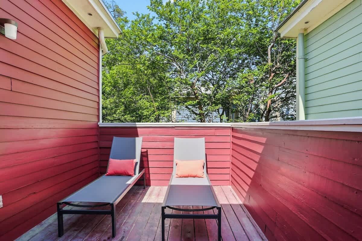 Una terraza de madera roja con dos sillones de descanso y una pequeña mesa, con vista a una zona verde bordeada de árboles.