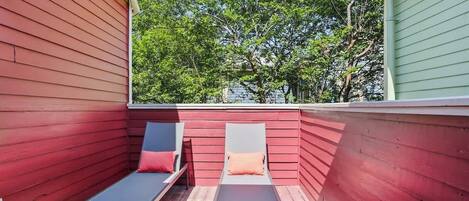 Una terraza de madera roja con dos sillones de descanso y una pequeña mesa, con vista a una zona verde bordeada de árboles.