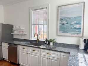 A modern kitchen with white cabinetry, a granite countertop, a stainless steel refrigerator, and a window with blinds.