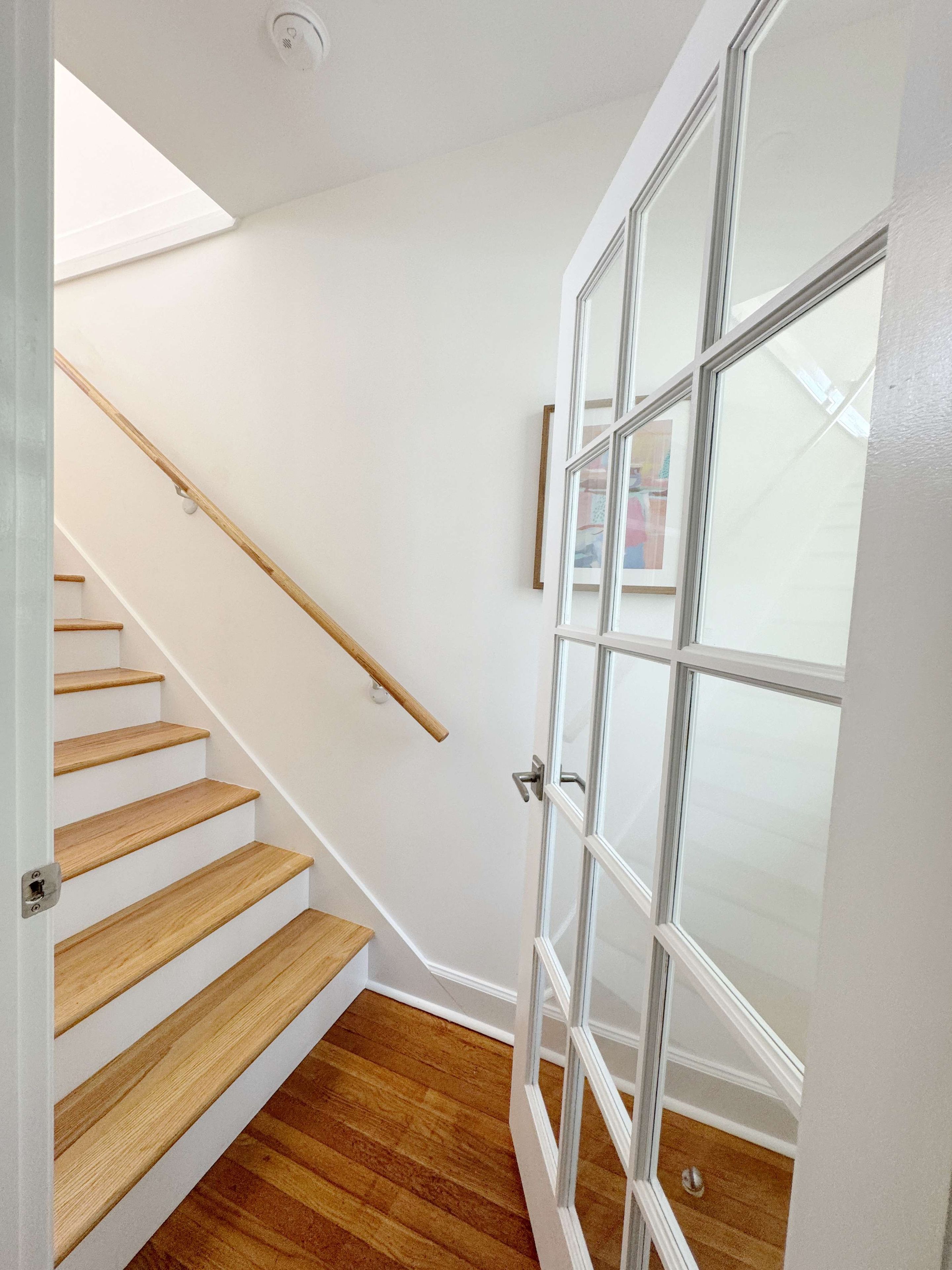 Una escalera de madera con peldaños que conducen a un rellano, una puerta de cristal y una lámpara en el techo.