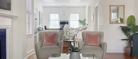 A spacious living room with a glass coffee table, two armchairs with cushions, a fireplace, and a view into the kitchen.