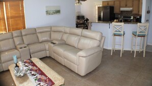 A living room with a beige sectional sofa, a coffee table with a decorative runner, and a kitchen area with wooden cabinets and bar stools. - Navarre Beach Escape-Pet & Family Friendly (Navarre)