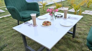 Une table à manger d’extérieur recouverte d’une nappe blanche, avec des chaises vertes et un vase rempli de fleurs roses.