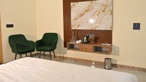 Une chambre d’hôtel avec un grand lit, deux fauteuils verts, une console en bois surmontée d’une œuvre d’art en marbre et une petite poubelle.