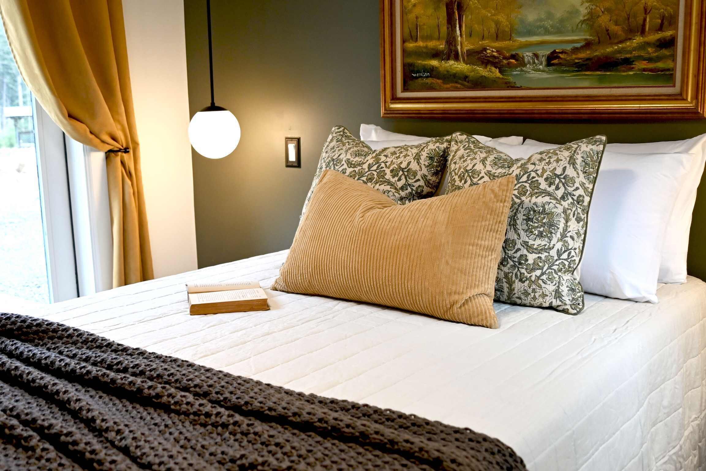 Una cama bien hecha con edredón blanco, varios almohadones y un libro sobre la cama.