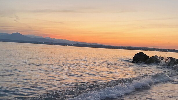 Ein Strand mit sanft rauschenden Wellen, einem klaren Himmel und fernen Bergen im Hintergrund.