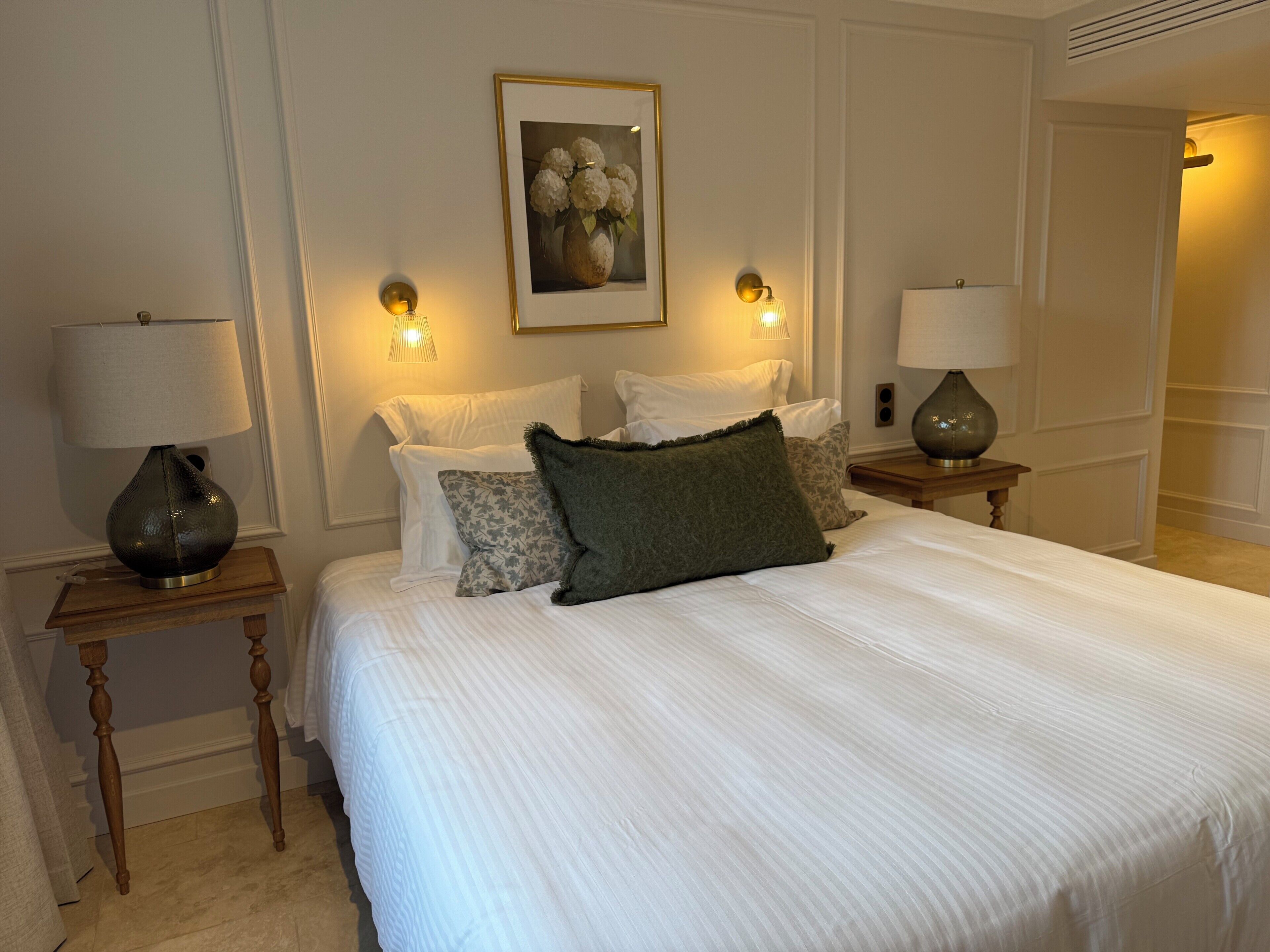Habitación de hotel ordenada con una cama grande, dos mesitas de noche con lámparas, una obra de arte enmarcada sobre la cama y una puerta que da a otra habitación.