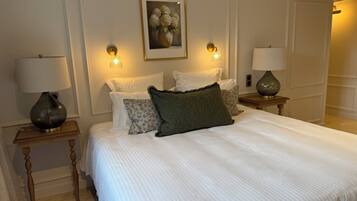 Habitación de hotel ordenada con una cama grande, dos mesitas de noche con lámparas, una obra de arte enmarcada sobre la cama y una puerta que da a otra habitación.