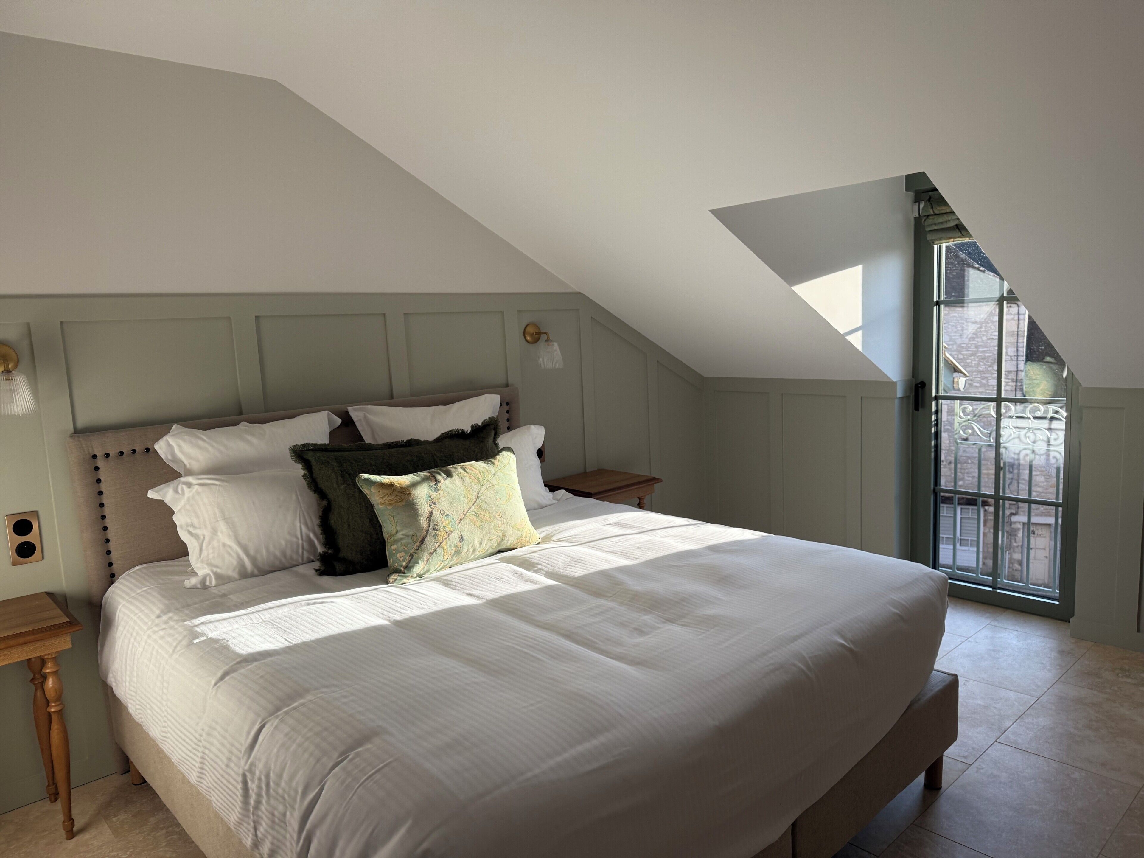 A bedroom with a sloped ceiling, a bed with multiple pillows, a nightstand, and a window with a view.