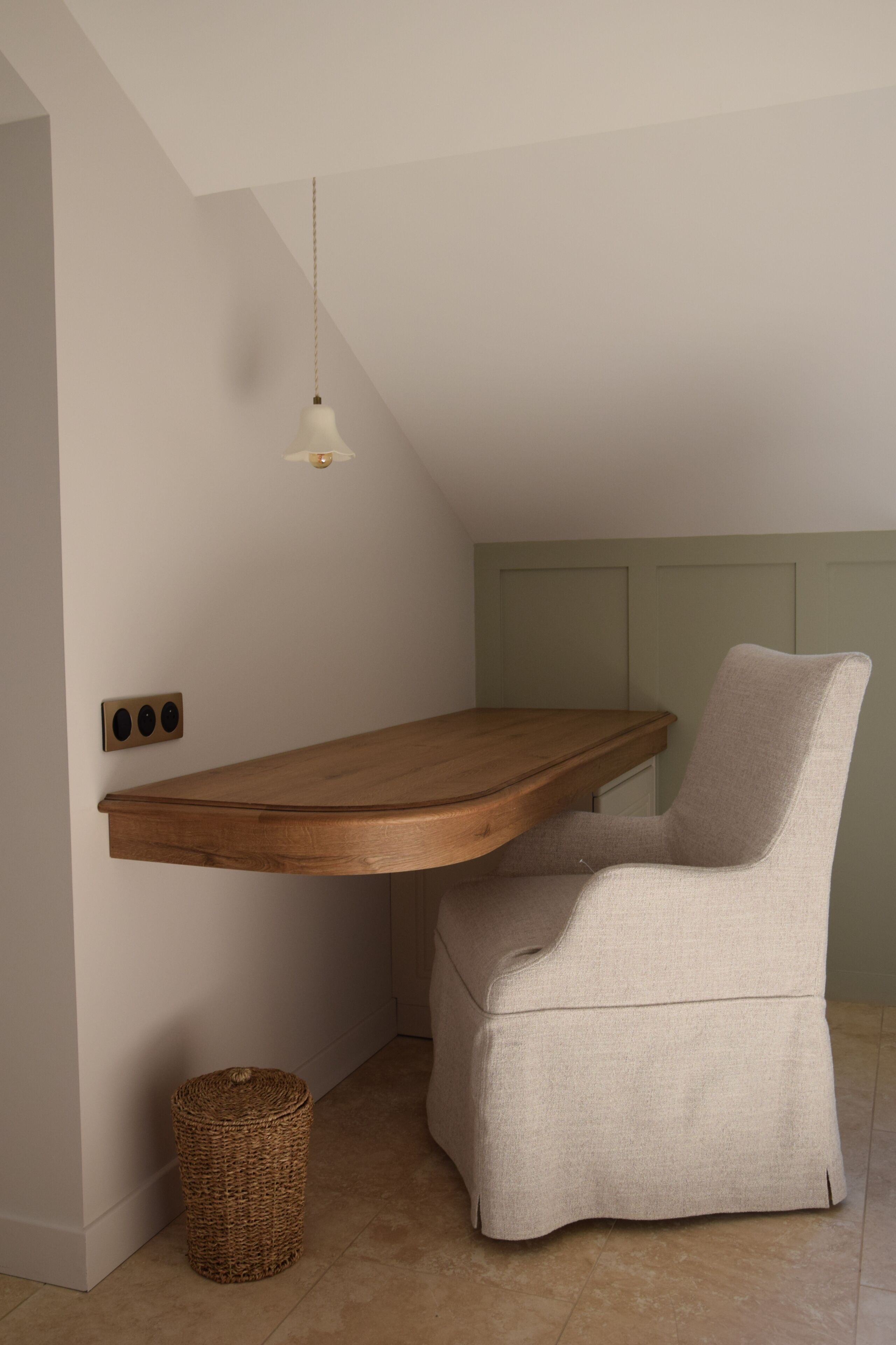 Una habitación de esquina con un escritorio de madera, una silla, una lámpara colgante y una cesta de mimbre.
