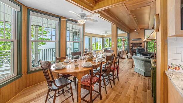Un coin repas avec une table en bois dressée pour six personnes, entourée de chaises, de grandes fenêtres et d’un ventilateur de plafond.