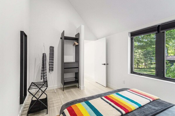 Un dormitorio moderno con un ventanal grande, una cama con manta a rayas, un estante de metal negro y una puerta que da a otra habitación.