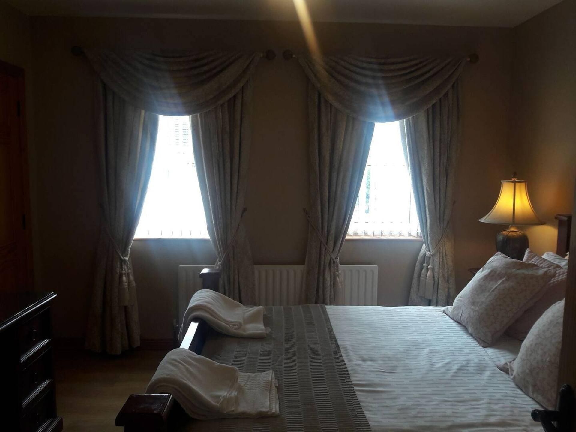 A hotel room with a bed, two windows with curtains, a bedside lamp, and a radiator.