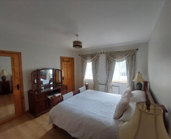 A bedroom with a large bed, a wooden dresser, a mirror, a lamp, and a window with curtains. - Carndonagh - 3-bedrooms - Sleeps 6/7 - Garden (Carndonagh)