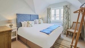 Un dormitorio con una cama grande, una mesita de noche, una silla de madera, una ventana con cortinas y un techo inclinado.