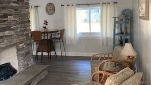 A living room with a stone fireplace, wicker furniture, a dining area with chairs, and a large window with curtains. - Looking for a beach escape? Find out what Costal Corner has to offer your family (Oak Island)