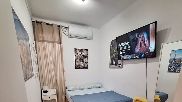 A compact bedroom with a bed, a TV mounted on the wall, a small bedside table, and a wooden shelf.