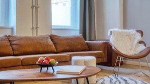 Una sala de estar moderna con un sofá de cuero marrón, una mesa de centro de madera con un cuenco de frutas y un libro, y una butaca blanca mullida.